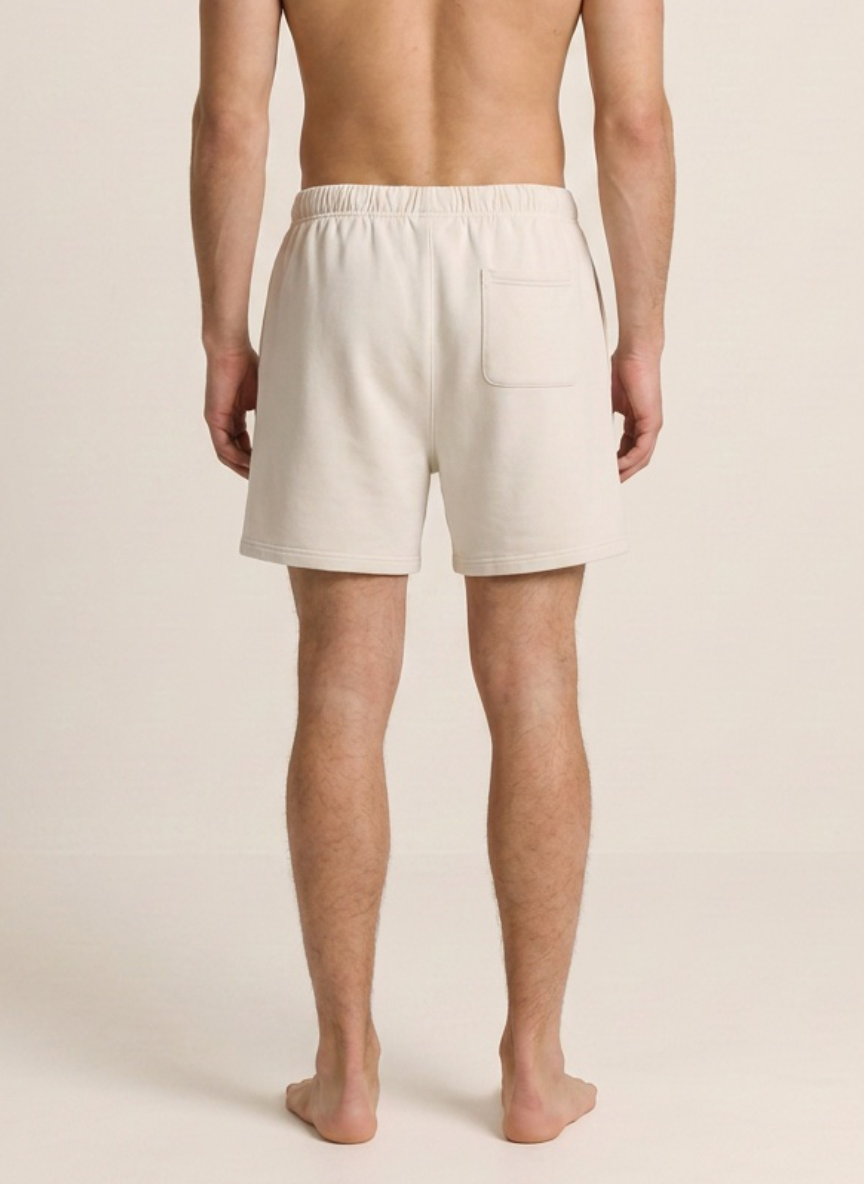 A man shown from the back wearing light cream-colored lounge shorts with an elastic waistband and a side pocket, standing barefoot against a plain beige studio background.