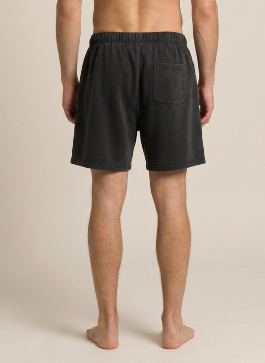 Rear view of a man wearing dark charcoal gray lounge shorts with an elastic waistband and a side pocket, standing barefoot against a light beige studio background.