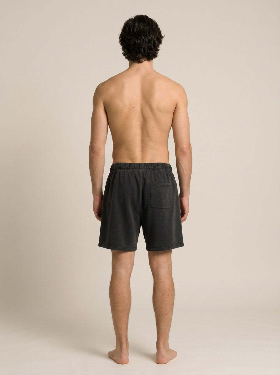 A man viewed from behind wearing dark charcoal gray lounge shorts, standing against a plain beige studio background.