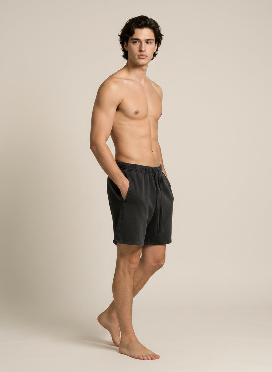 A shirtless male model wearing dark charcoal gray drawstring shorts, standing in profile against a plain beige studio background.