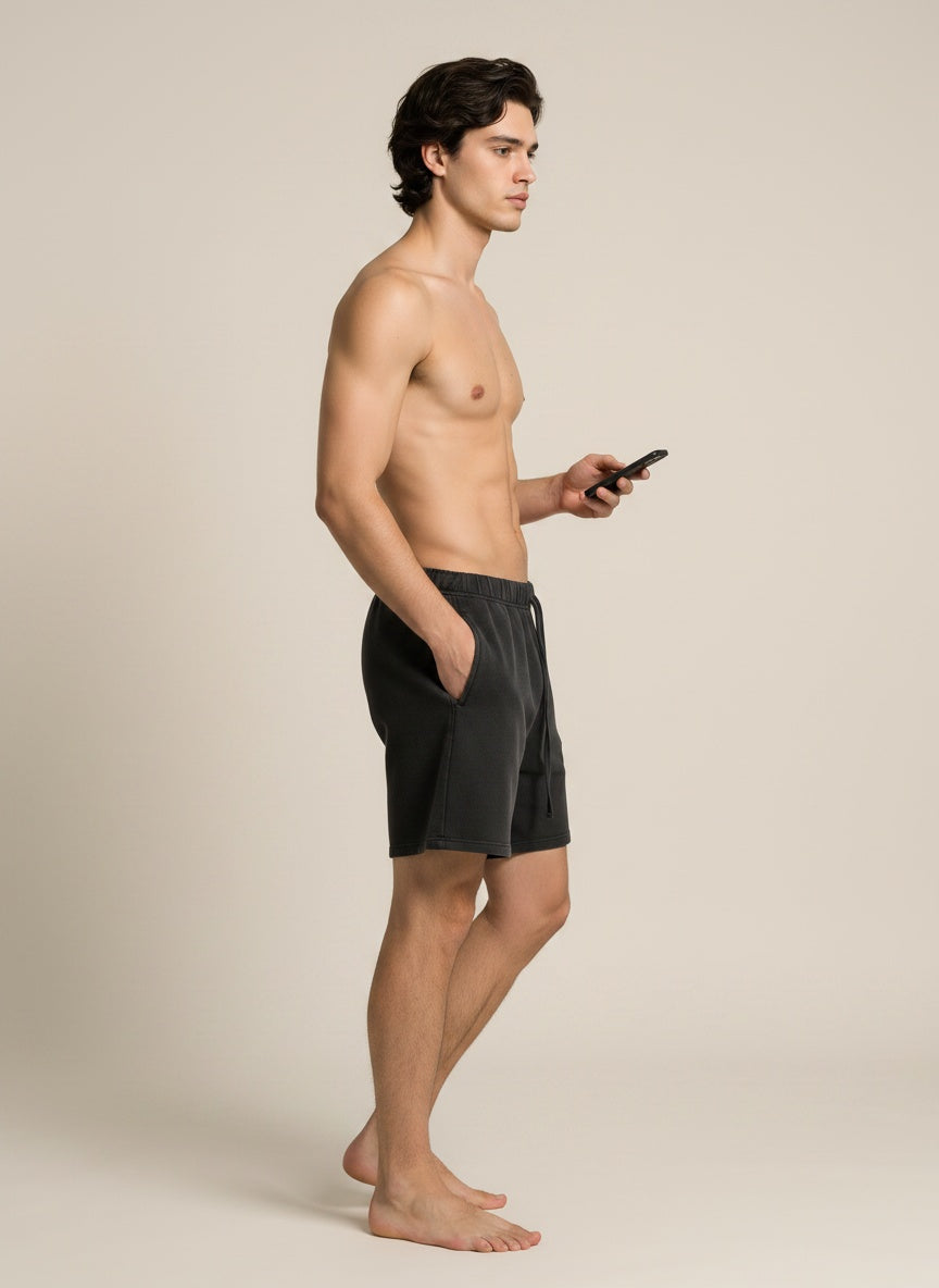 A shirtless man standing in profile wearing dark charcoal gray swim trunks, holding a black smartphone against a plain warm beige studio background.