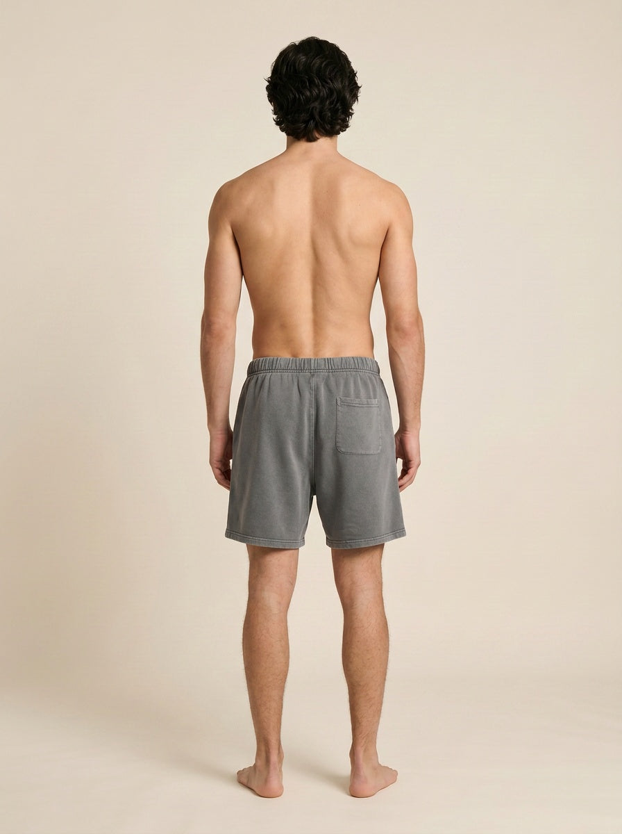 A man viewed from behind wearing gray lounge shorts, standing barefoot against a plain beige studio background.