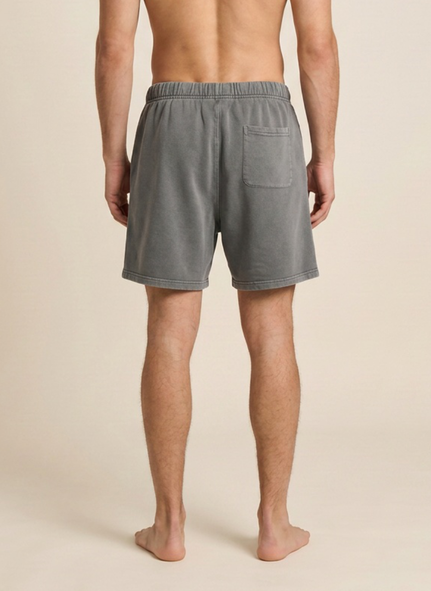 A man viewed from the back wearing gray lounge shorts, standing barefoot against a plain beige studio background.