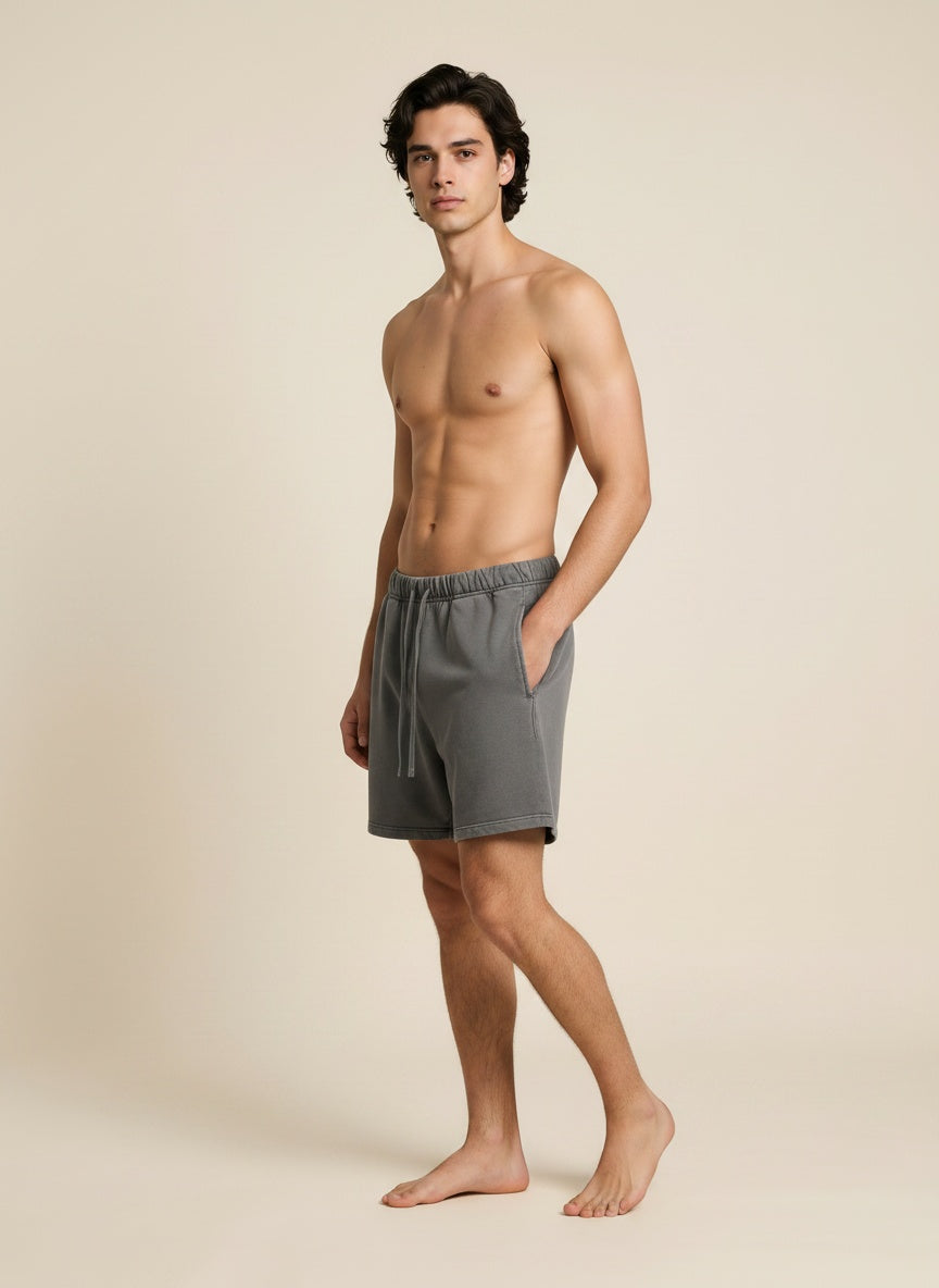 A shirtless man modeling gray drawstring shorts against a plain beige studio background.