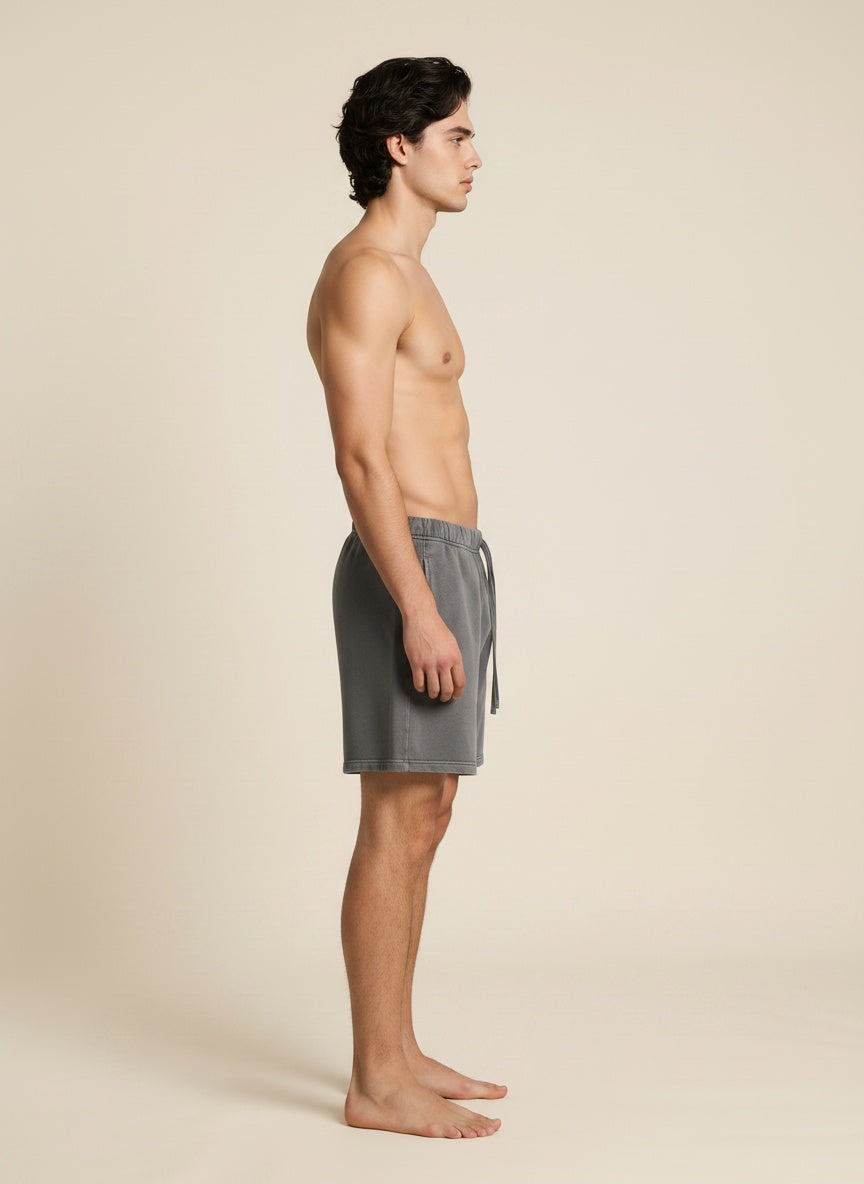 A man in side profile wearing gray drawstring shorts, standing barefoot against a plain beige studio background.