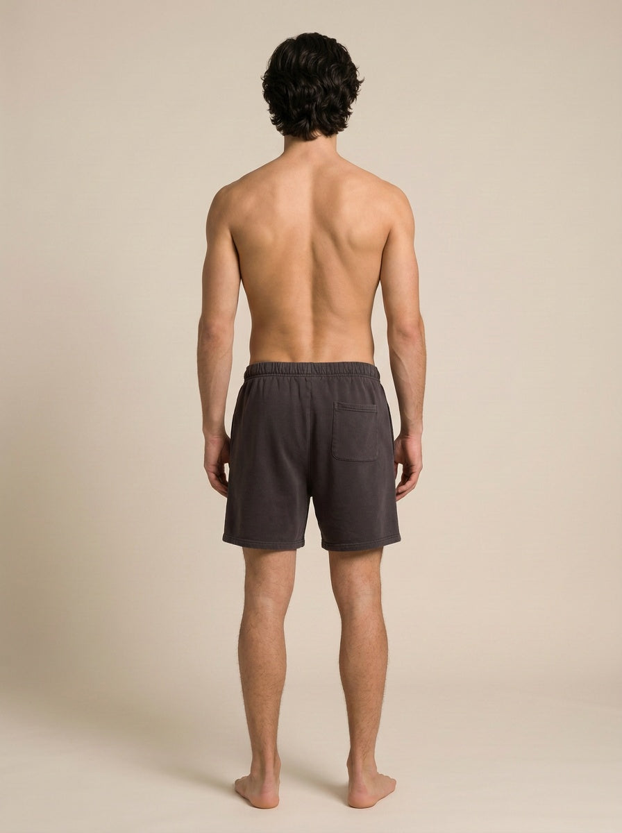 A man viewed from behind wearing dark purple lounge shorts, standing barefoot against a solid beige studio background.
