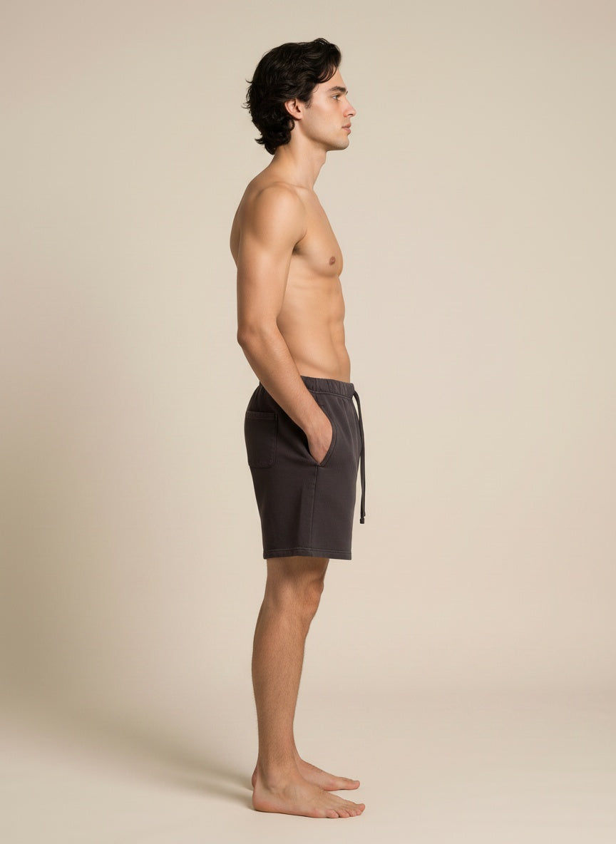 A man stands in profile wearing dark charcoal gray drawstring shorts against a plain beige studio background.