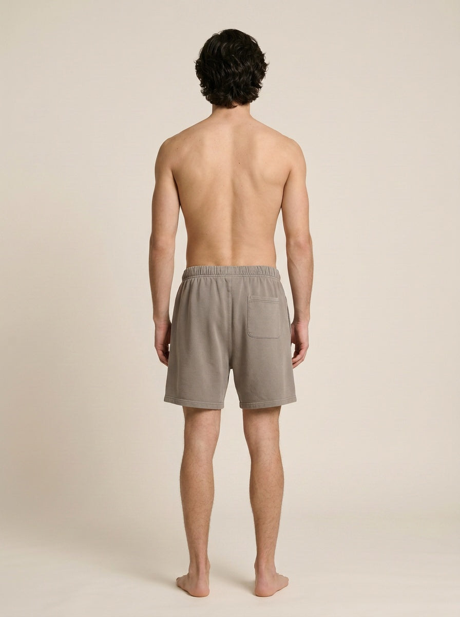A man viewed from behind wearing light taupe-gray drawstring shorts with a side pocket, standing against a plain beige studio background.