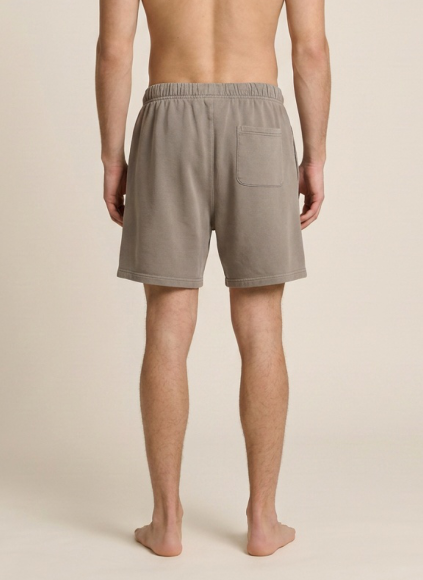 A man viewed from the back wearing light taupe-gray drawstring shorts with a side pocket, standing barefoot against a plain beige studio background.