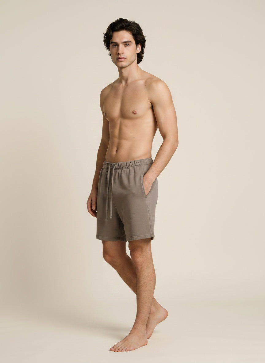 A shirtless male model wearing taupe drawstring shorts, standing against a solid beige studio background.