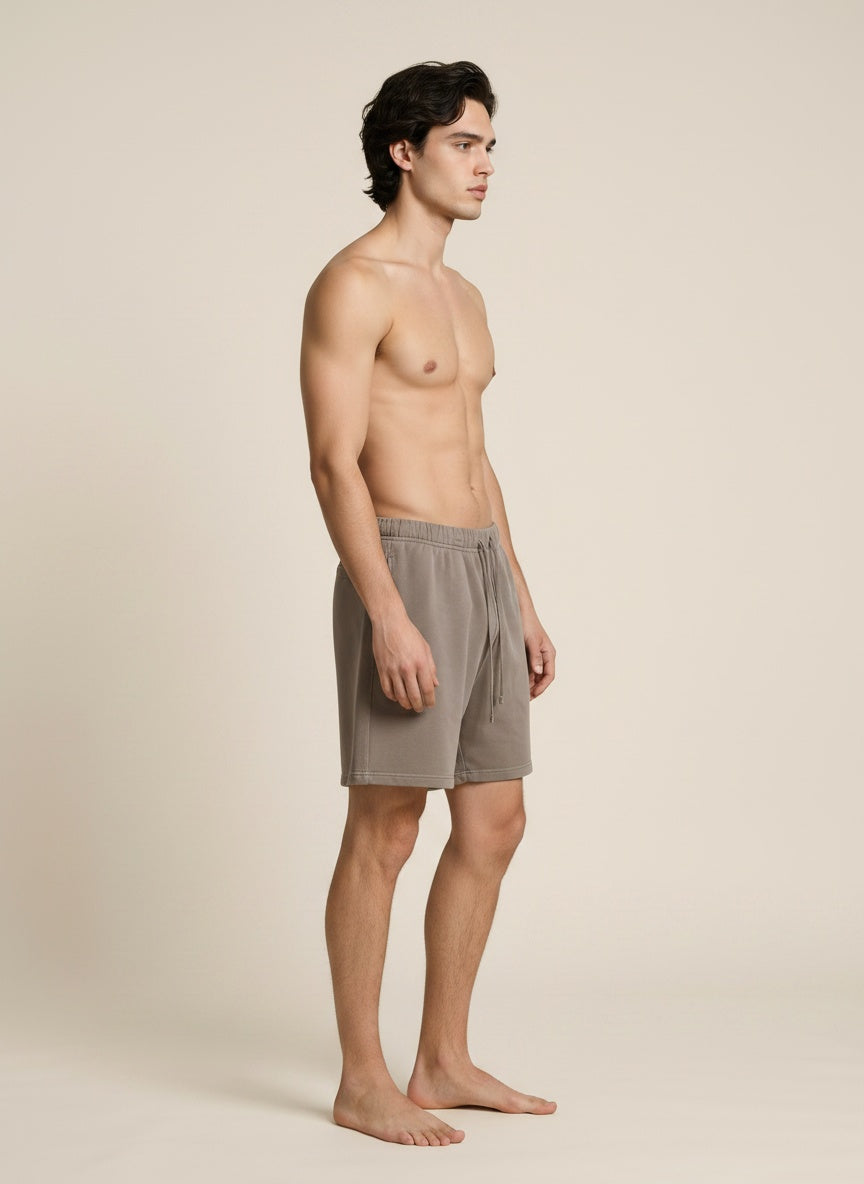 A man stands in profile wearing light taupe drawstring shorts against a plain warm beige studio background.