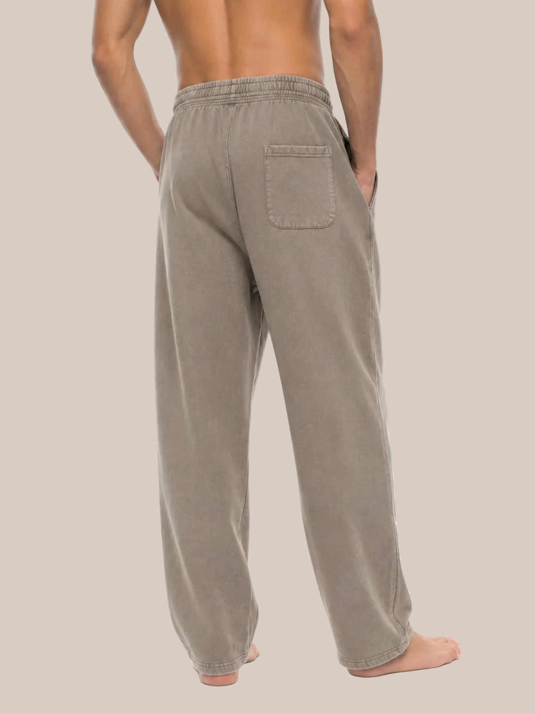 A man shown from the back wearing loose, wide-leg lounge pants in a warm taupe or light brown color, standing barefoot against a plain beige background.