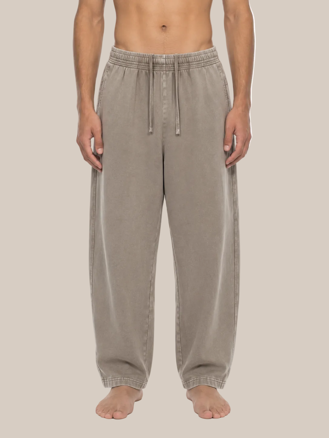 A person modeling a pair of loose, wide-leg lounge pants in a light taupe-beige color, shown from the waist down against a plain white background.