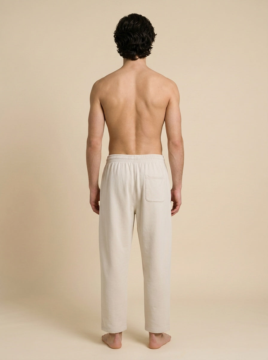 A man shown from the back wearing light beige lounge pants, standing barefoot against a solid beige studio background.