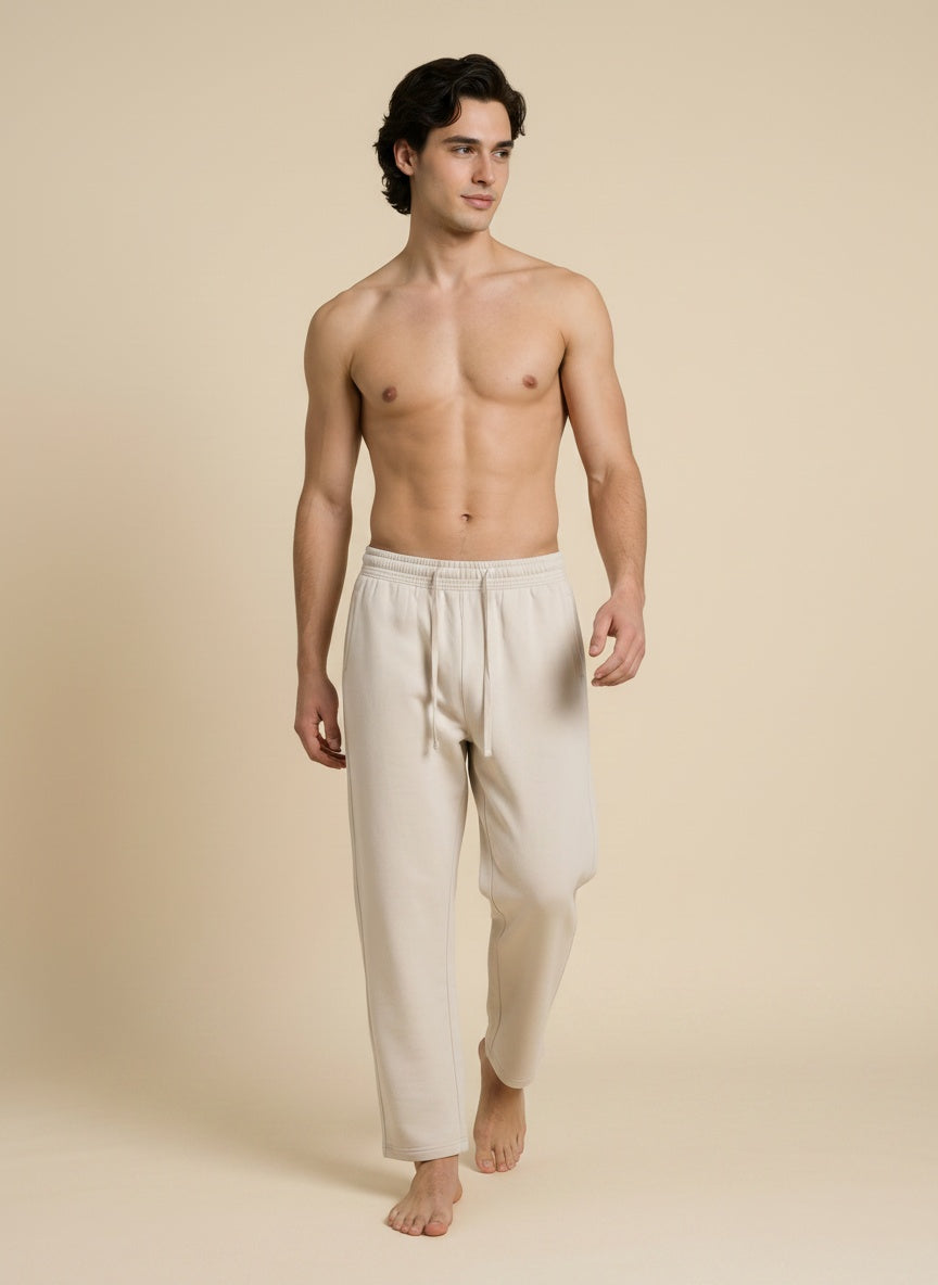 A shirtless male model wearing light beige drawstring lounge pants, standing against a solid beige studio background.