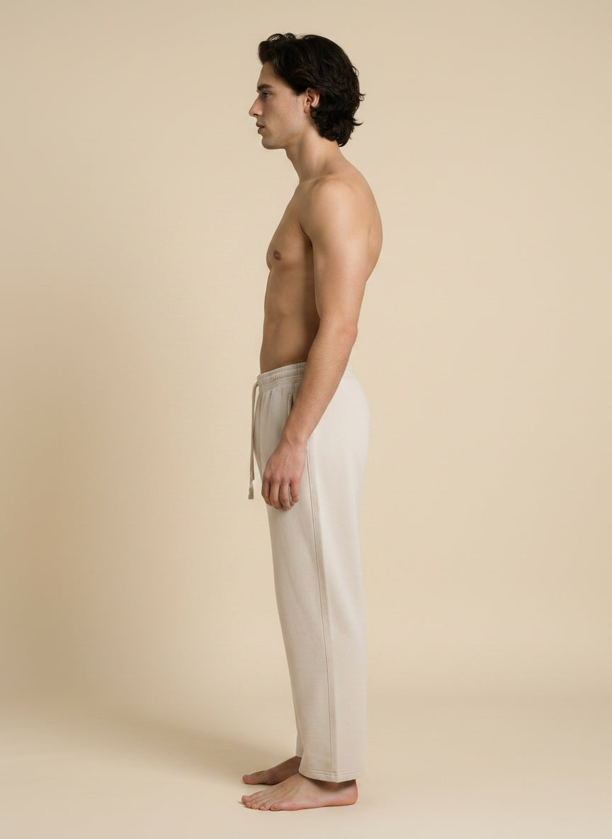 A man in side profile wearing light beige drawstring lounge pants, standing barefoot against a solid beige studio background.