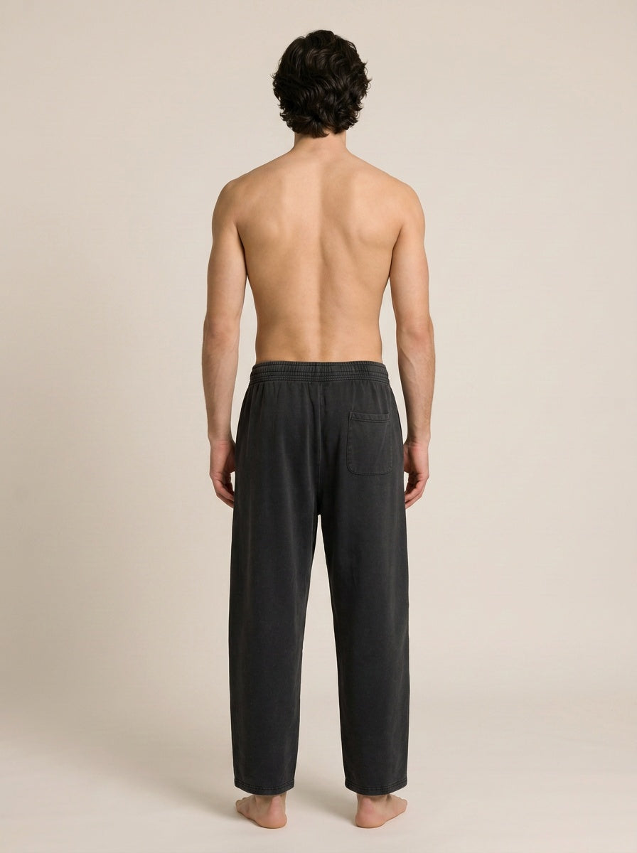 A man viewed from behind wearing dark charcoal gray lounge pants, standing barefoot against a plain beige studio background.