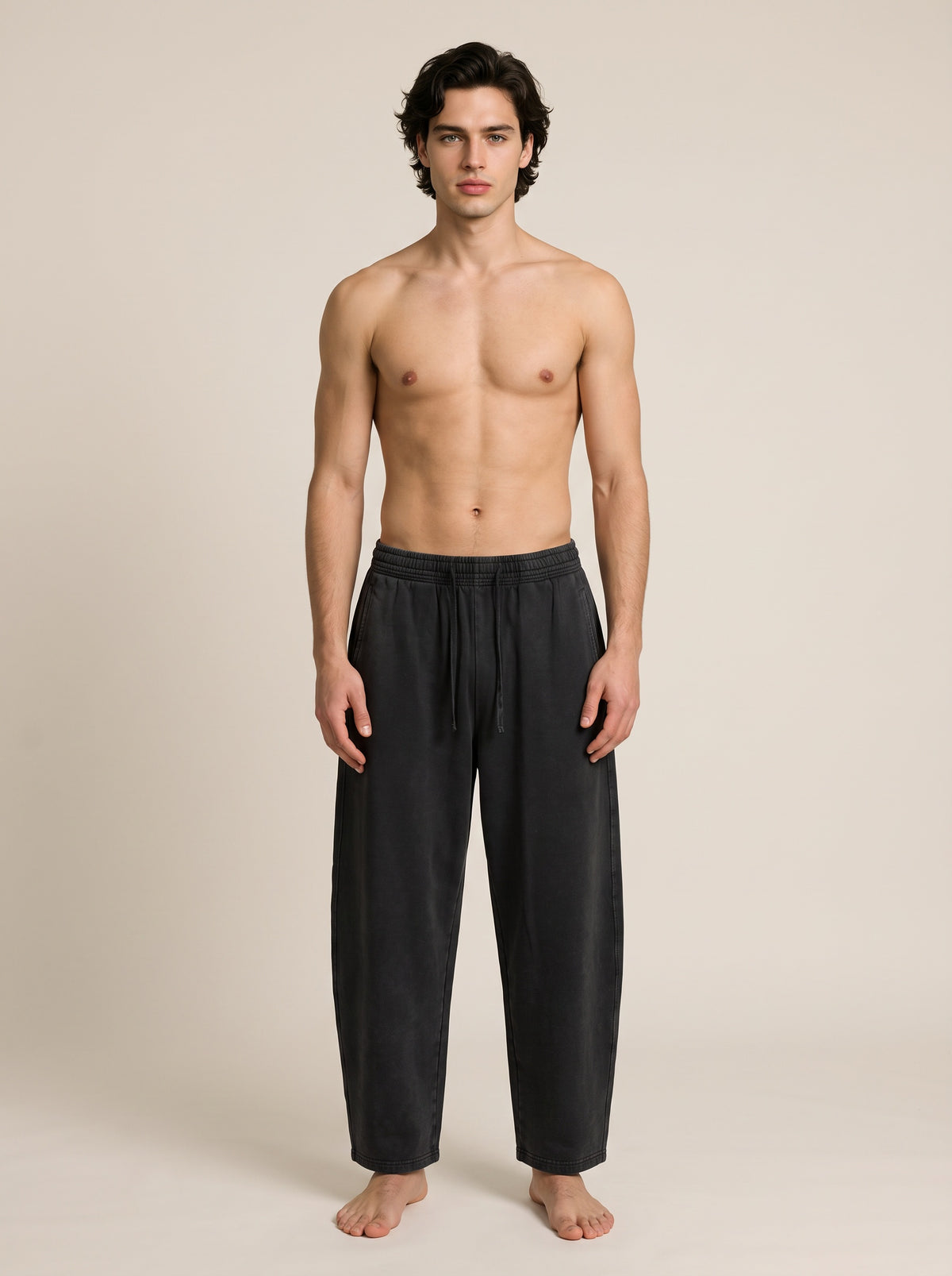 A shirtless male model stands barefoot against a plain warm beige studio background, wearing loose dark charcoal lounge pants with a drawstring waist.