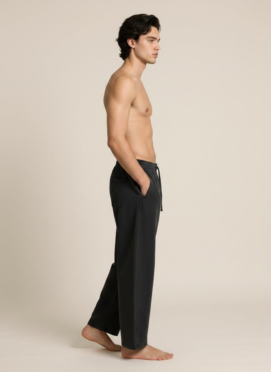 A shirtless man stands in profile wearing loose black drawstring pants against a plain warm beige studio background.