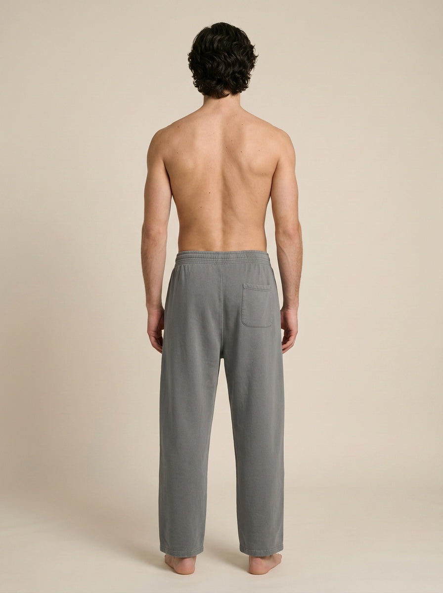 A man viewed from behind wearing gray lounge pants, standing barefoot against a plain beige studio background.