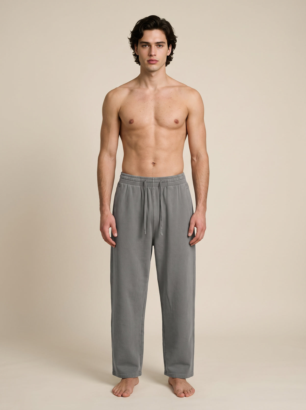 A shirtless male model stands barefoot against a plain beige studio background, wearing loose gray drawstring lounge pants.