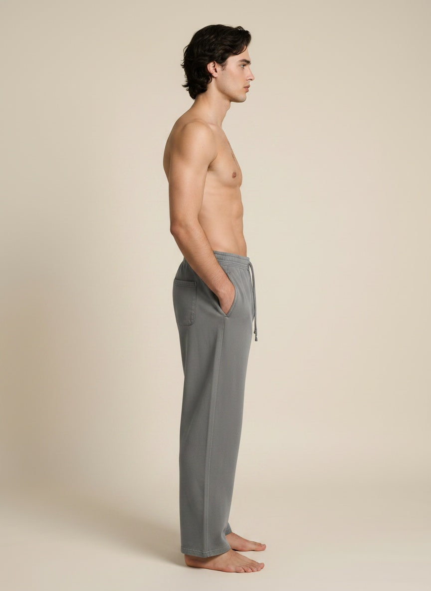 A man in side profile wearing gray drawstring lounge pants, standing barefoot against a plain beige studio background.