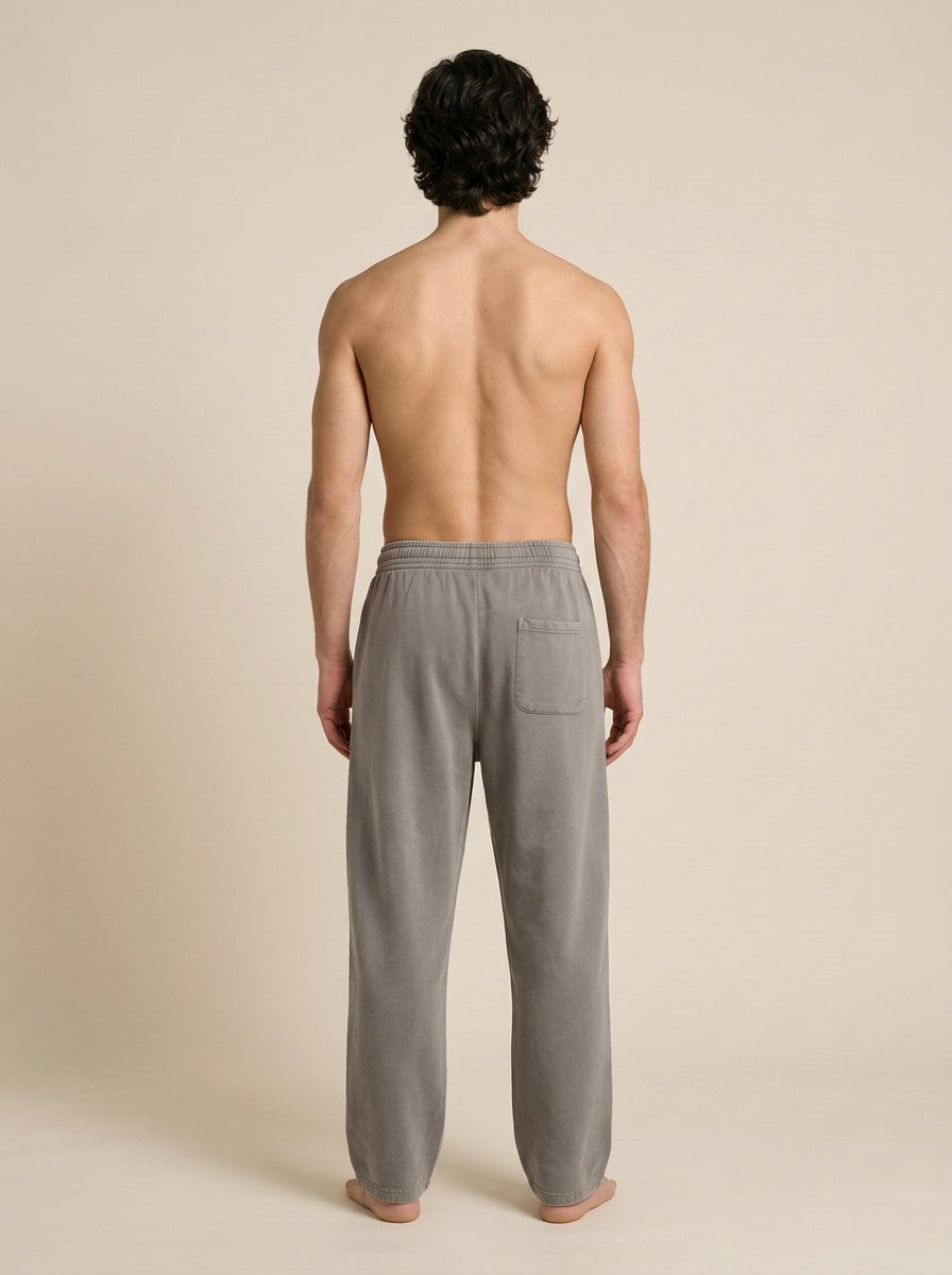 A man viewed from behind wearing light gray lounge pants, standing barefoot against a plain beige studio background.
