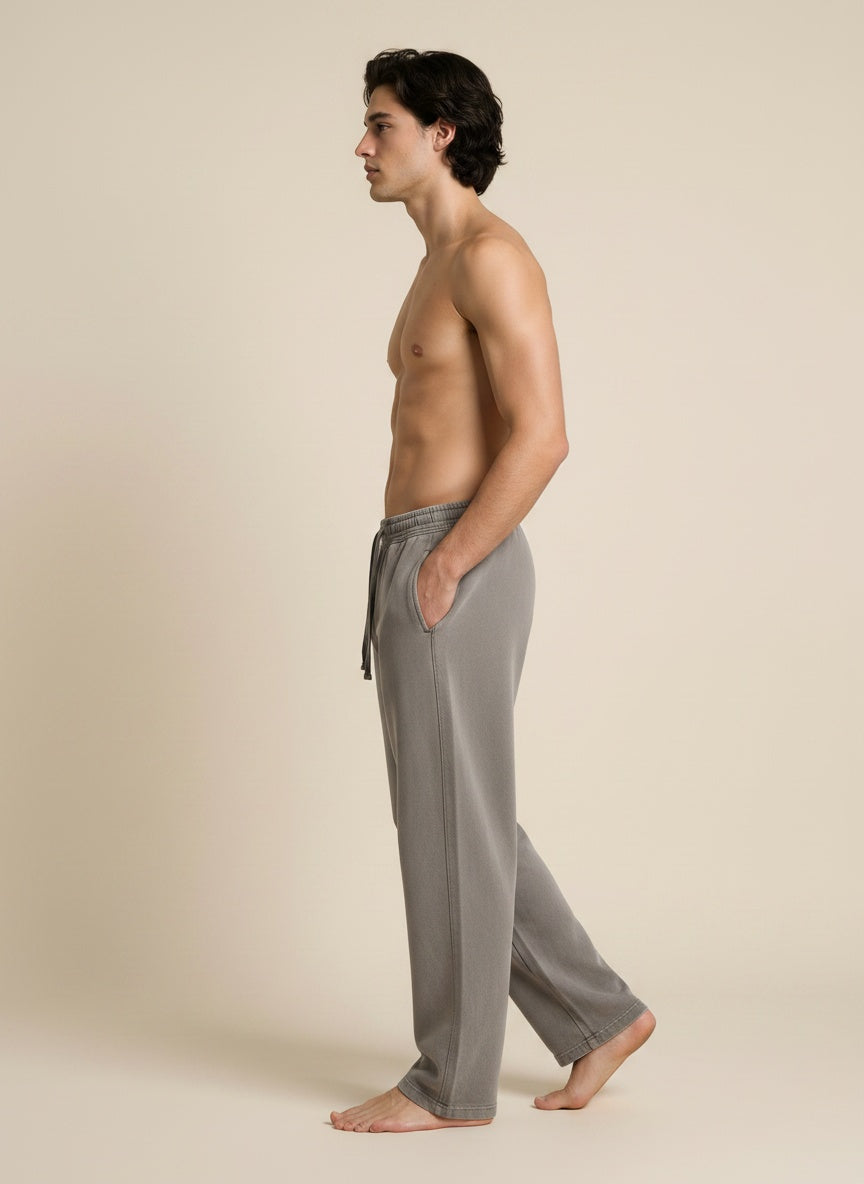 A man in side profile wearing light gray drawstring lounge pants, standing barefoot against a plain beige studio background.