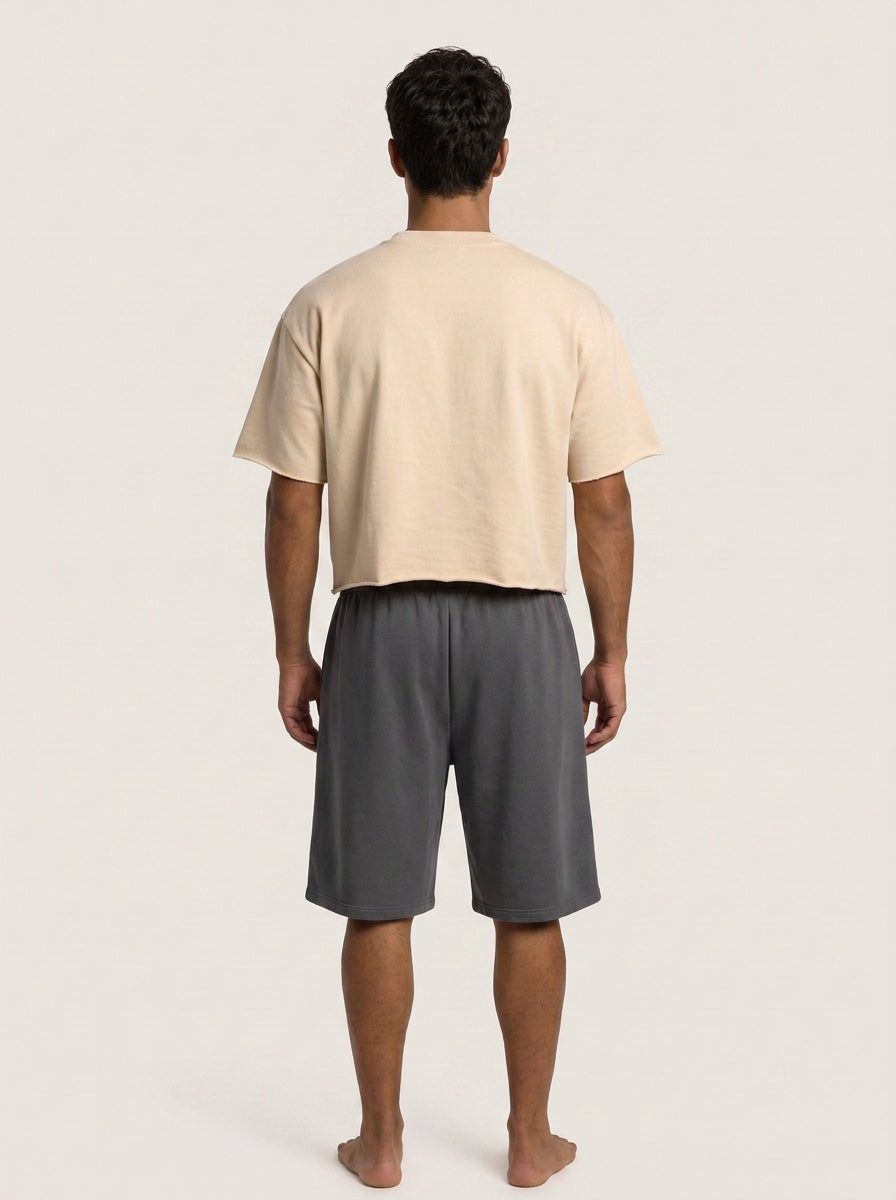 A man viewed from behind wearing a light beige cropped t-shirt and dark gray shorts against a plain light gray studio background.