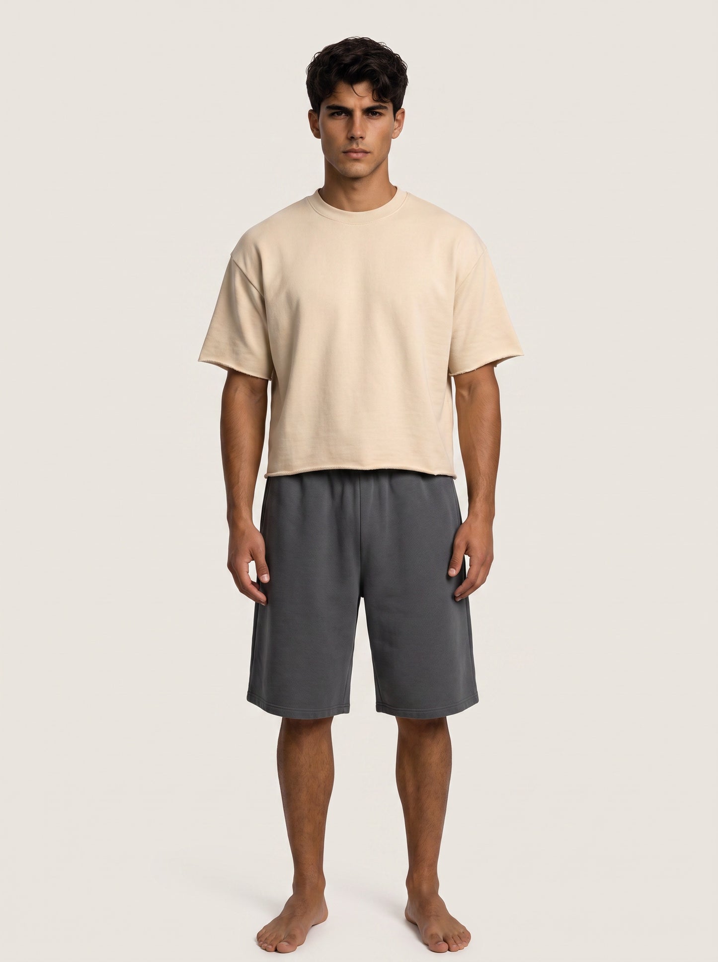 A man stands barefoot against a plain light beige studio background, wearing a loose short-sleeve cream t-shirt and dark gray shorts.