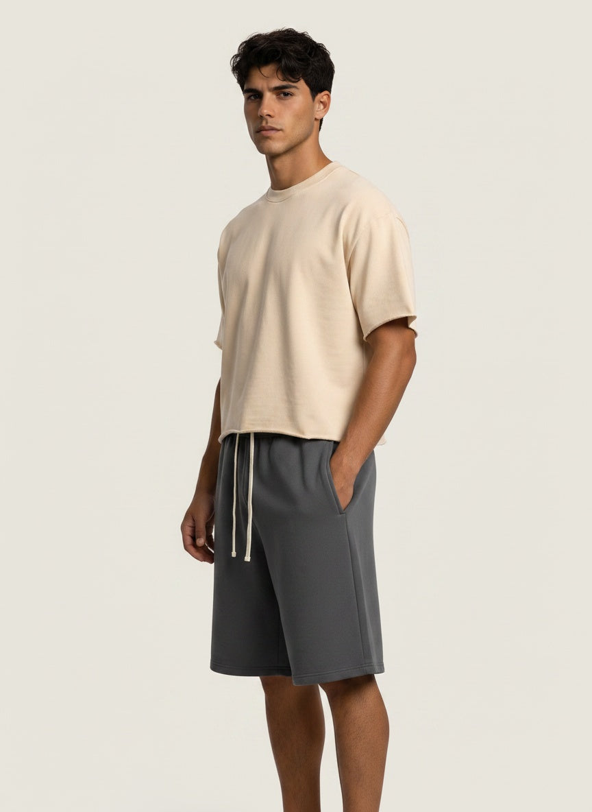 A man modeling a light beige short-sleeve crewneck t-shirt and dark gray drawstring shorts against a solid off-white background.