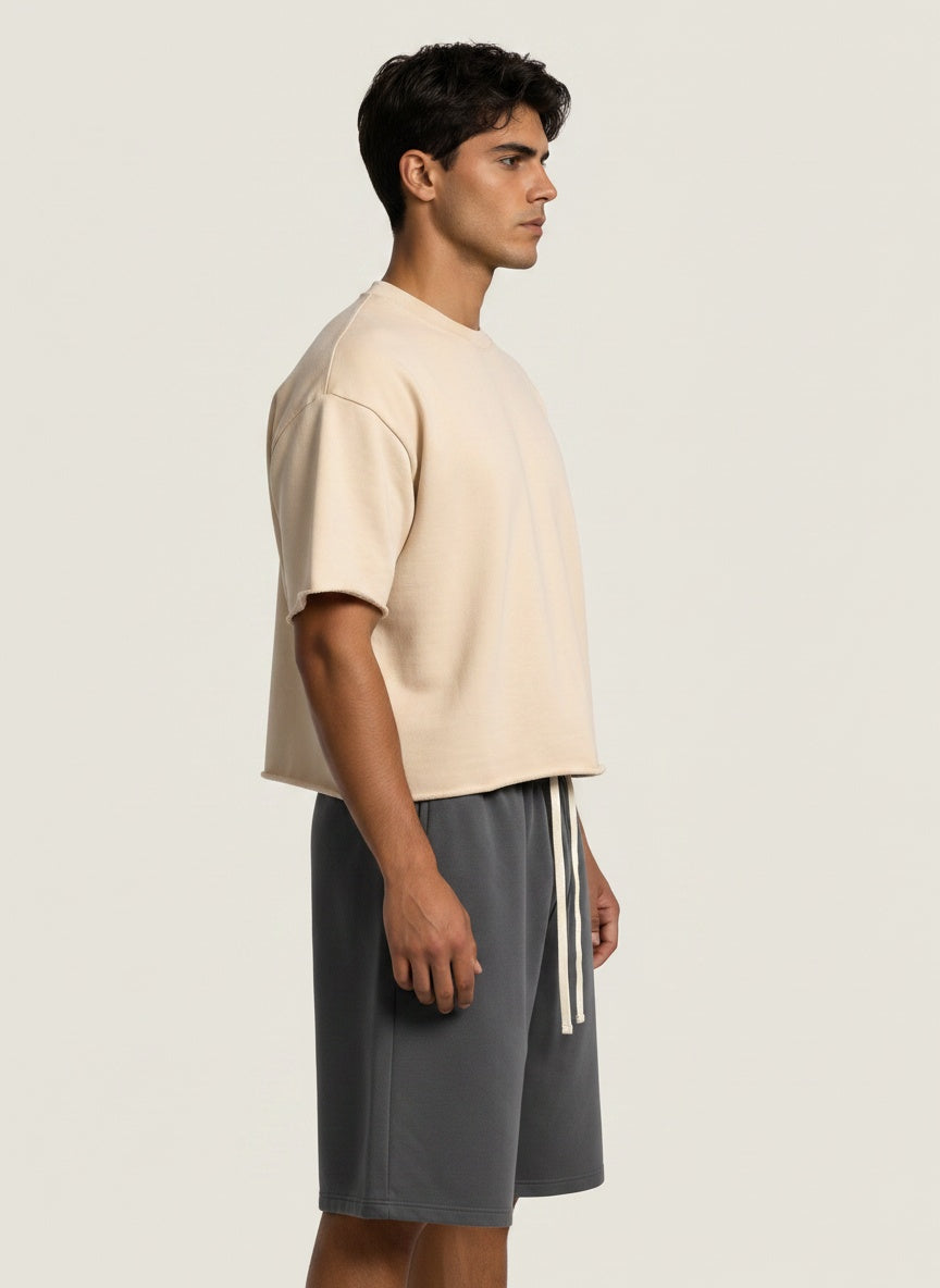 A man in profile wearing a light beige short-sleeve knit top and dark gray drawstring shorts against a plain light beige studio background.