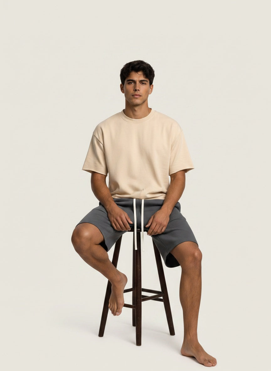 A man sits on a dark wooden stool wearing a light beige crewneck t-shirt and dark gray shorts with white drawstrings, against a solid off-white background.