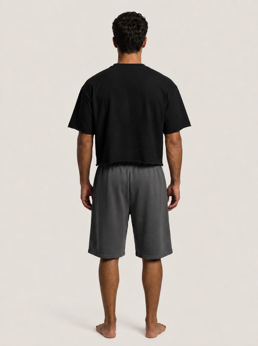 A man viewed from the back wearing a black short-sleeve t-shirt and dark gray shorts, standing barefoot against a light beige studio background.