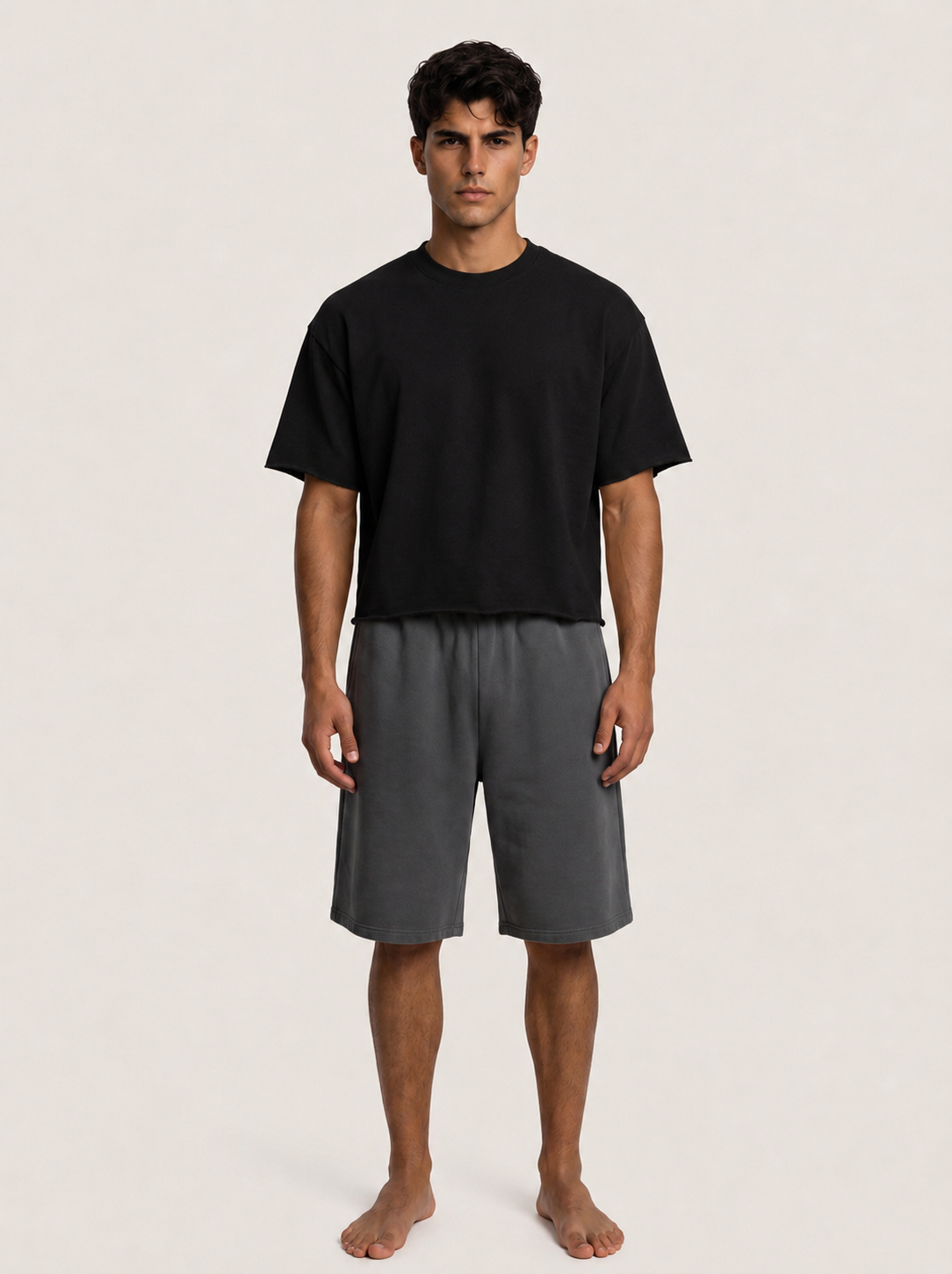 A man stands barefoot against a plain light beige studio background, wearing a black short-sleeve crew-neck T-shirt and dark gray drawstring shorts.