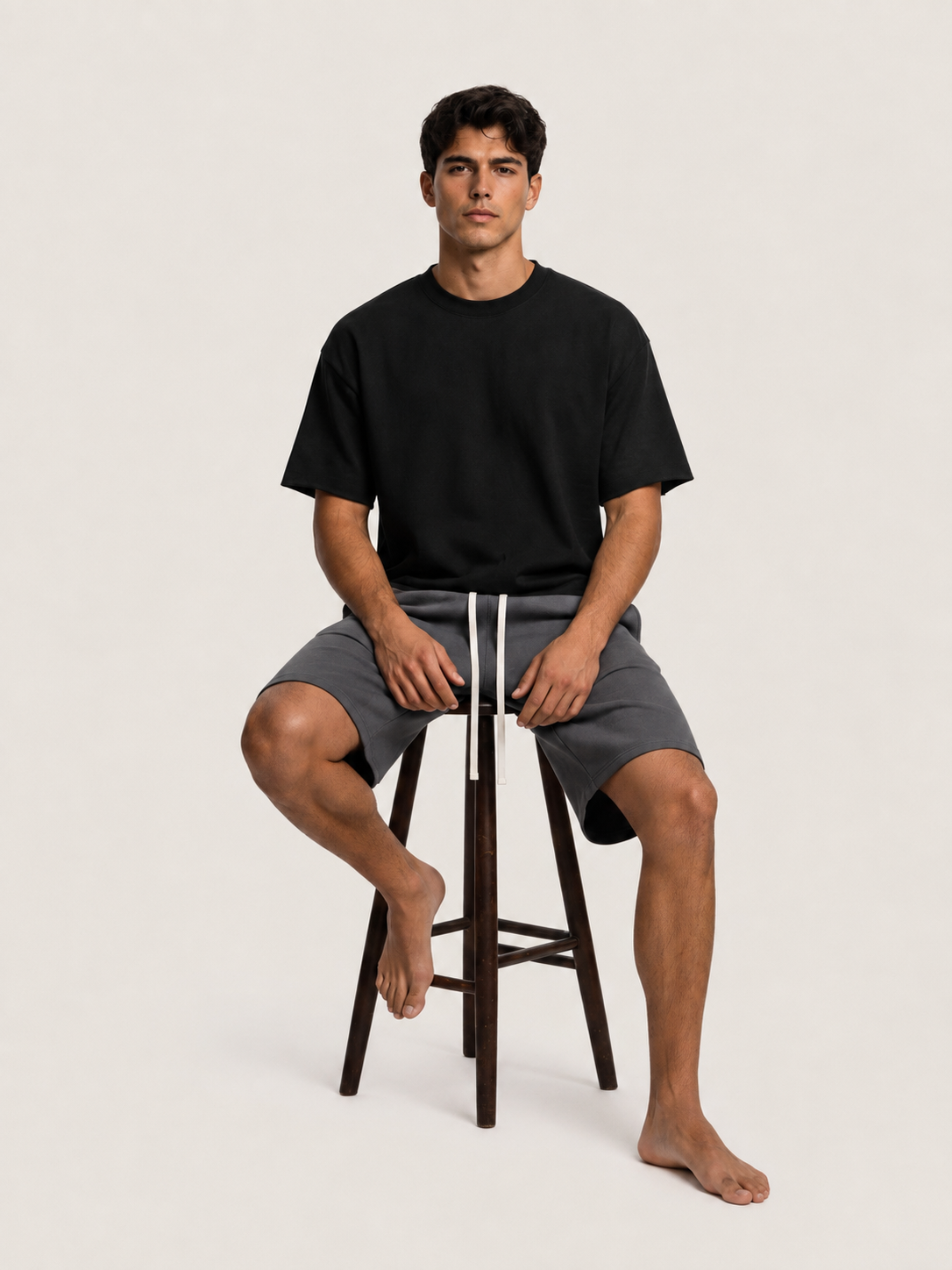 A man sits on a dark wooden stool wearing a black short-sleeve crewneck t-shirt and gray shorts with a white drawstring, against a solid light beige background.