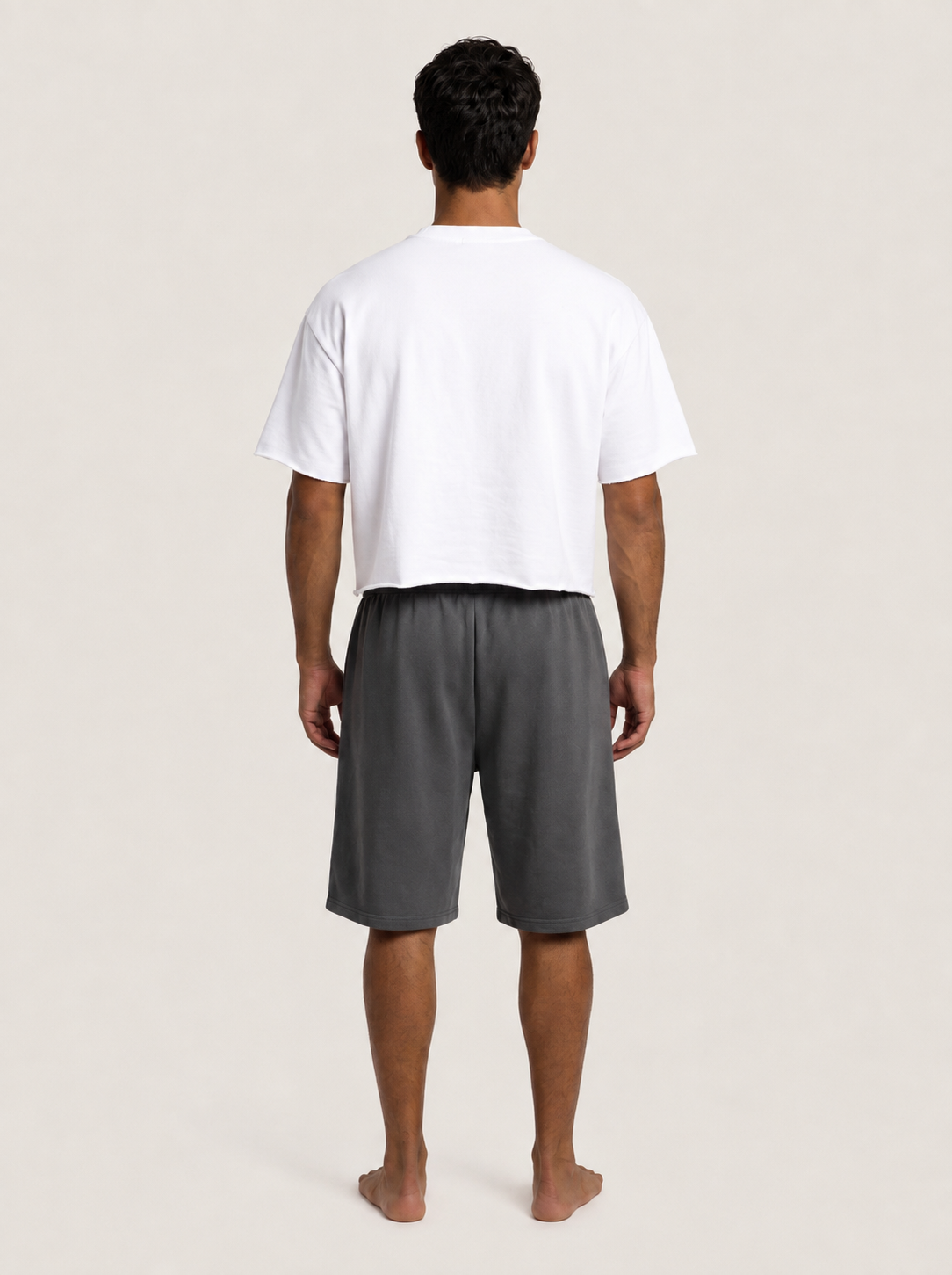 A man viewed from behind wearing a white cropped t-shirt and dark gray shorts, standing barefoot against a light beige studio background.