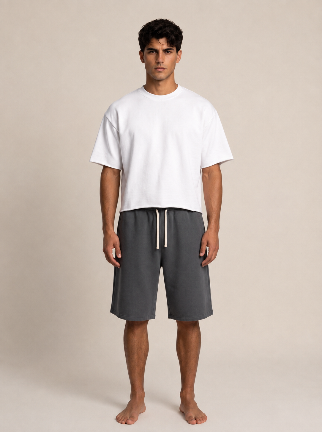 A man stands barefoot against a plain beige studio background, wearing an oversized white short-sleeve T-shirt and dark gray drawstring shorts.
