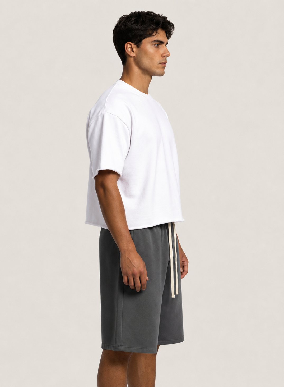 A man in profile wearing a white short-sleeve crewneck t-shirt and dark gray drawstring shorts, standing against a light beige studio background.