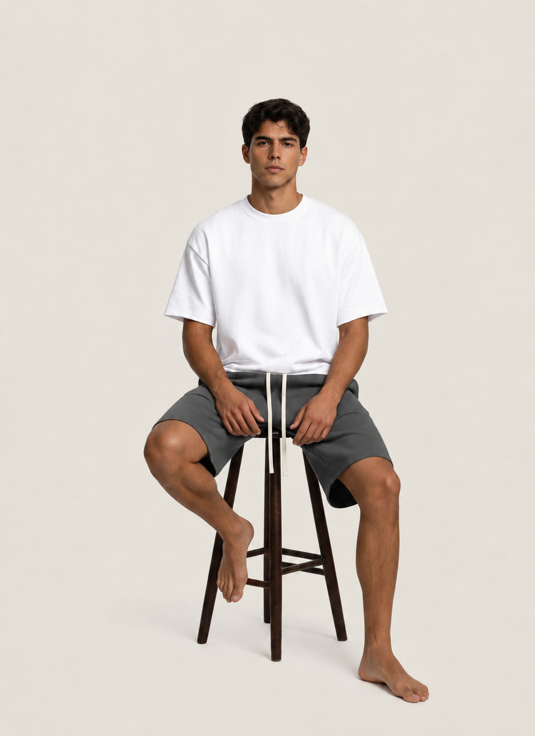 A man sits on a dark wooden stool wearing a white crewneck t-shirt and dark gray drawstring shorts against a solid light beige background.