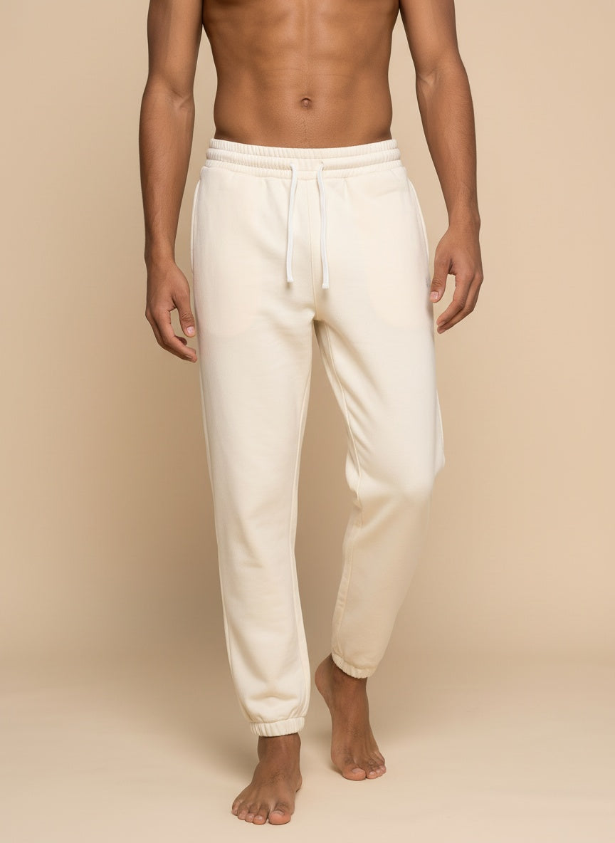 A man wearing off-white fleece jogger sweatpants with a drawstring waist and elastic cuffs, standing barefoot against a solid beige background.